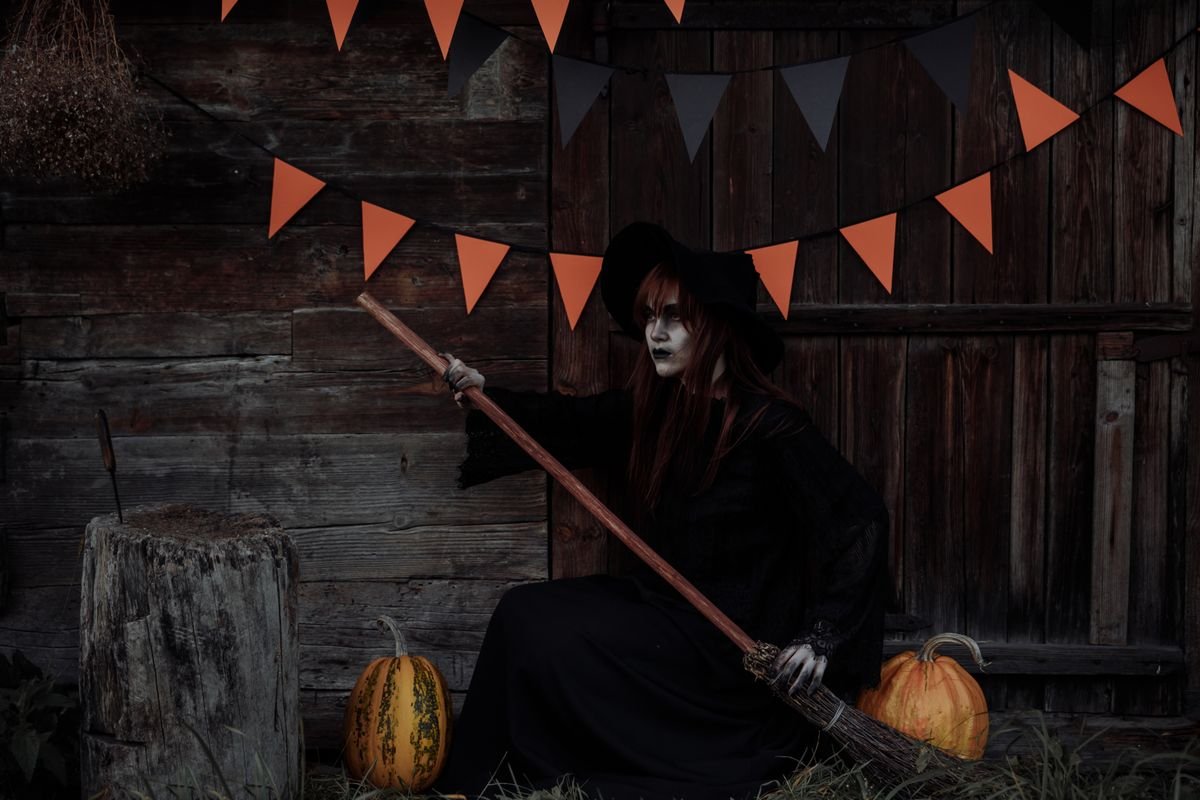 Witch seated with broom, pumpkins and orange bunting by a wooden shed